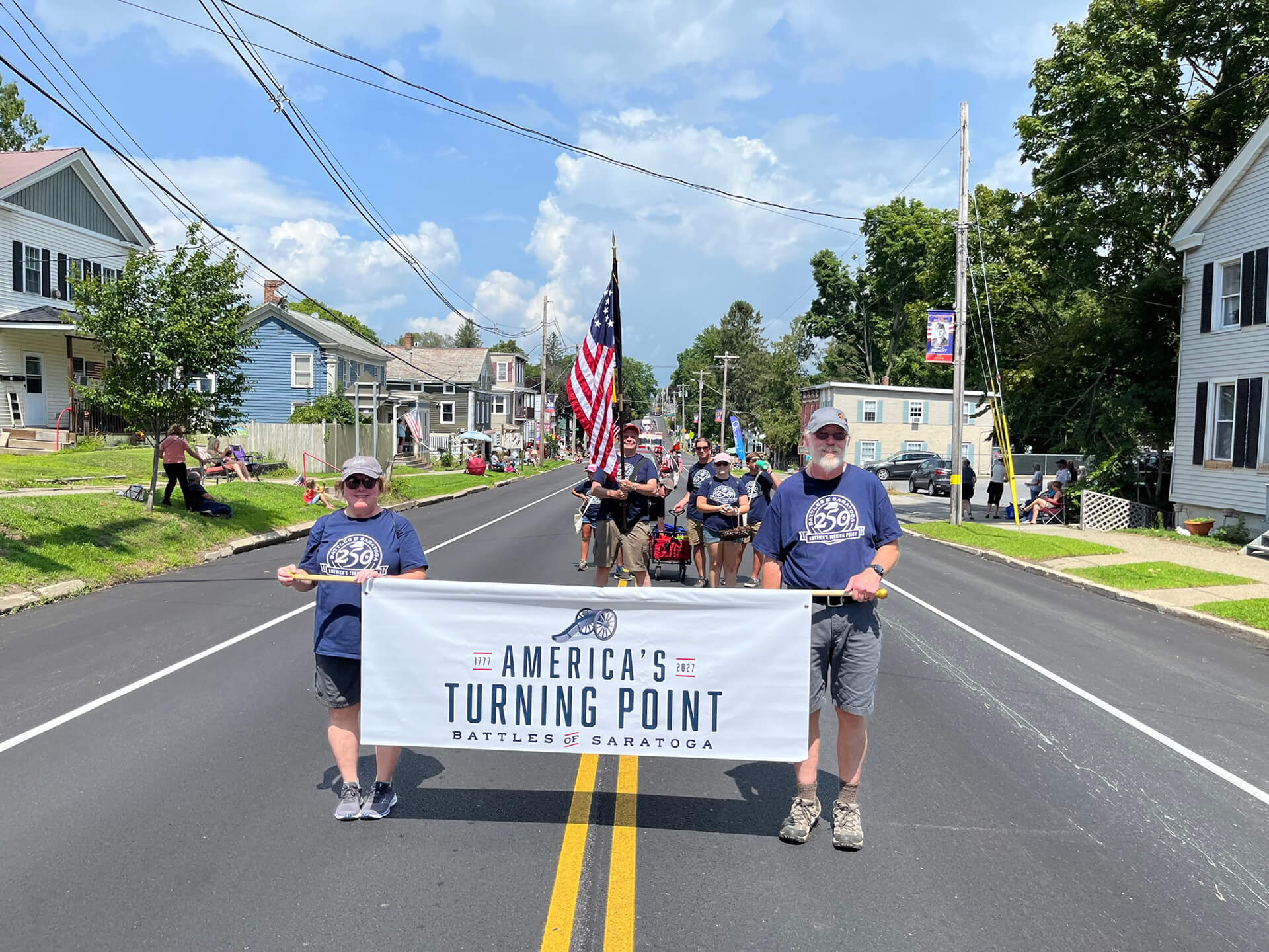 Saratoga 250 volunteers marching in a parade holding the American flag and a sign that reads "America's Turning Point - Battles of Saratoga (1777-2027)"