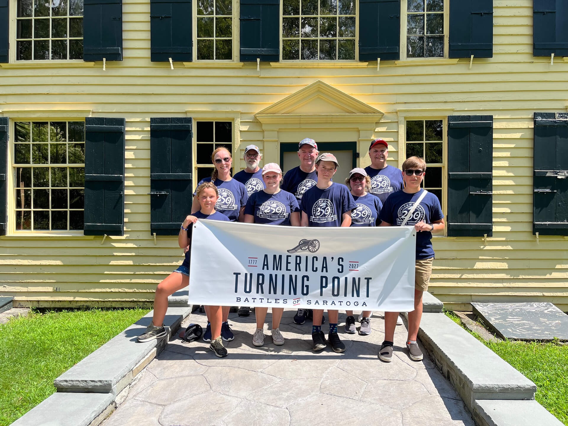 Saratoga 250 volunteers in front of The Schuyler House with a sign that reads "America's Turning Point - Battles of Saratoga (1777-2027)"