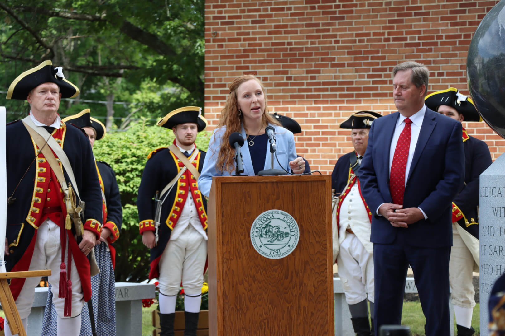 Woman speaking at a podium during a press conference with Revolutionary War reenactors in the background