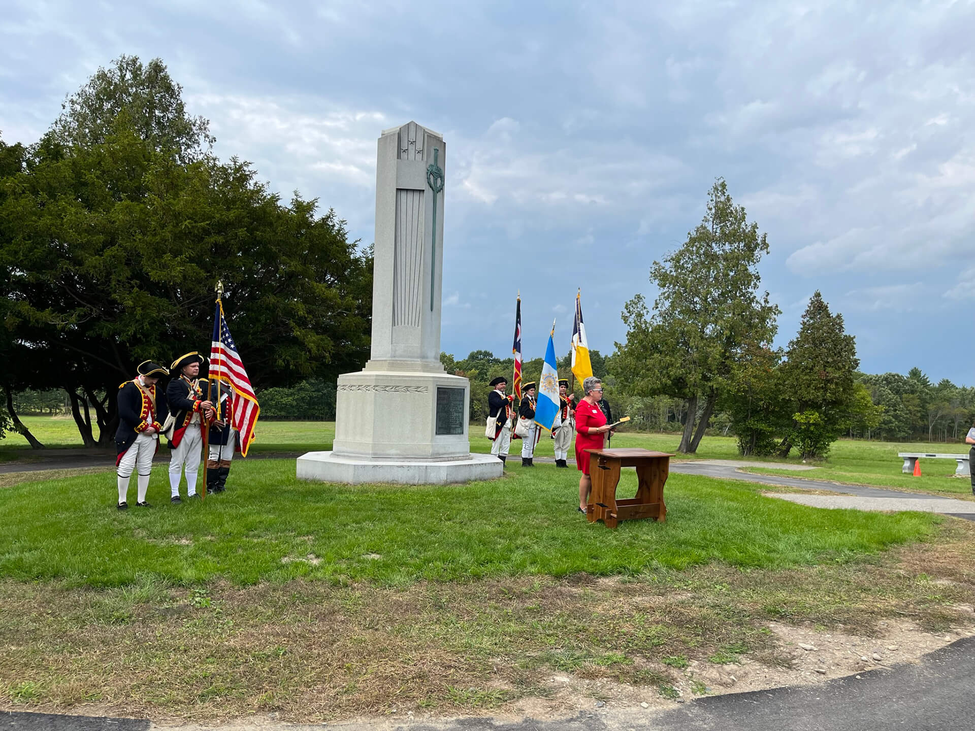 Revolutionary War reenactors with flags near a monument as a woman speaks at a podium