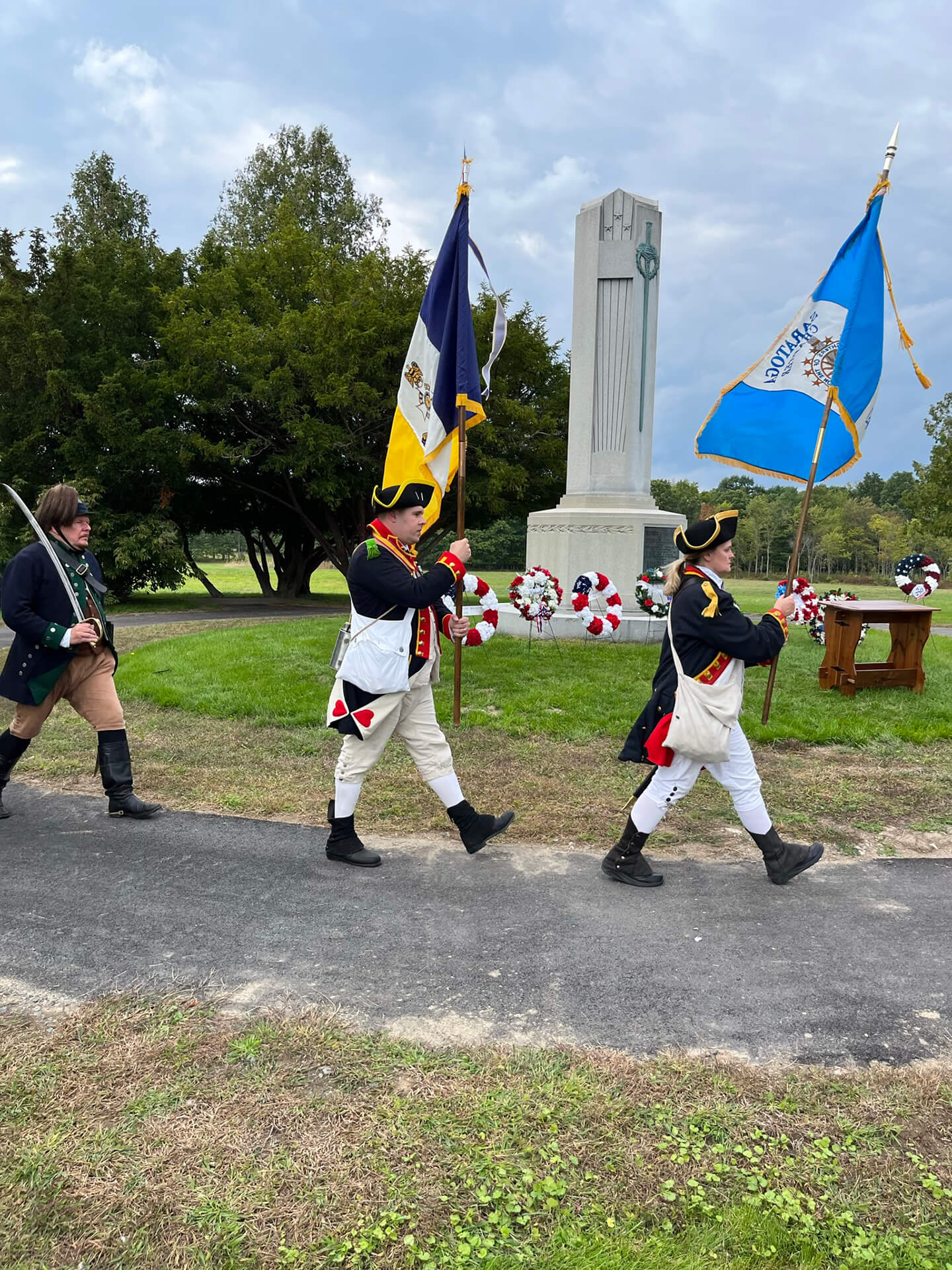 Three Revolutionary War reenactors marching with flags and swords near a monument