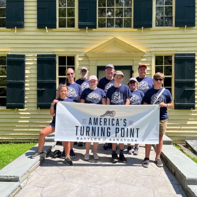 Saratoga 250 volunteers in front of The Schuyler House with a sign that reads "America's Turning Point - Battles of Saratoga (1777-2027)"