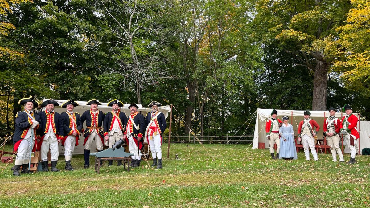 Woman speaking at a podium during a press conference with Revolutionary War reenactors in the background